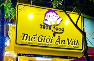 Trang Chủ 45 Bảng hiệu TOTO Food nền vàng, logo nhân vật và chữ nổi Thế Giới Ăn Vặt màu đen hắt sáng vào ban đêm